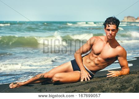 Summer. Muscular guy lying on his side. By the sea. Sculptural body. A beautiful young Italian man with a fabulous body lying on the sand by the sea. Abdominal showy.