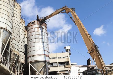 Ground level view of long demolition crane hand stretched up dismantling old metal tanks against blue sky