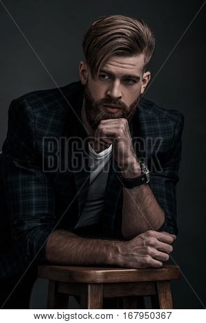 Stylishly confidence. Handsome young man in casual clothing looking away while leaning on stool against grey background