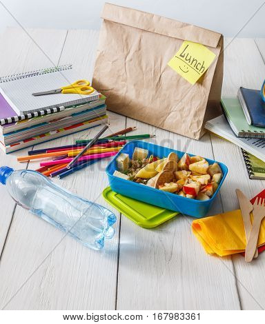 Healthy school lunch for child or teenager. Craft paper package, pile of exercise books, water, bag and food in lunch box on white wood table, cracker with cheese, nuts, oatmeal porridge and apples
