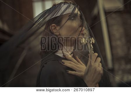 Portrait Of Praying Young Nun In Black Cassock, Cloak And Transparent Cape With Cross In Her Hands. 