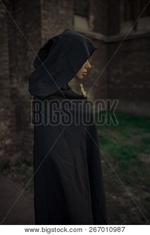 Beautiful Young Nun In Black Cassock And Hooded Cloak Is Standing And Praying With Cross In Her Hand