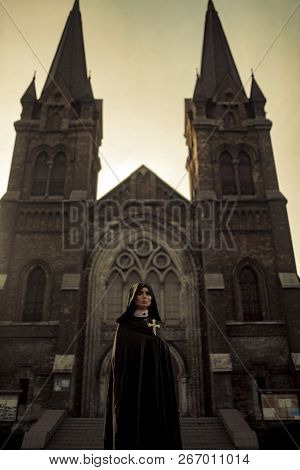 Beautiful Young Nun In Black Cassock And Hooded Cloak Is Standing With Cross In Her Hands On Temple 