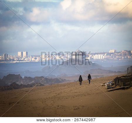 Two Women Walk In The Sand Towards The Senhor Da Pedra Chapel. Beach, Ocean, Back View, Cloudy Day.