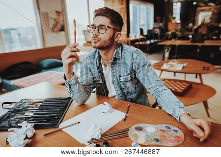 Painter Holds Paintbrush Looking Sits At Desk. Portrait Of Talanted Young Caucasian Handsome Man Pai