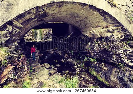 Tourist Woman Standing Under Arch Bridge And Little Waterfall In The Background, Norway. Traveling A