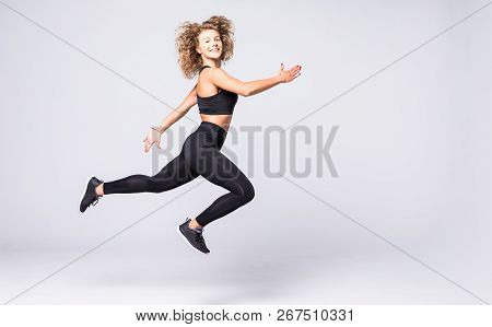 Beautiful Young Fit Woman With Curly Hair, Happily Jumping On A Grey Background. Slim Woman In Sport