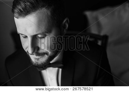 Stylish Groom In Black Suit And Bow Tie, Sitting In Chair At Window Light. Confident And Happy Portr