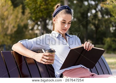 Woman Sitting On A Wooden Bench, Reading A Book And Drinking Coffee