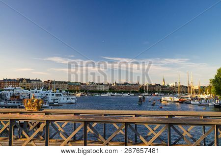 Golden Gilded Crown On Skeppsholmsbron Bridge Wooden Railing With View Of Ostermalm District With Tr