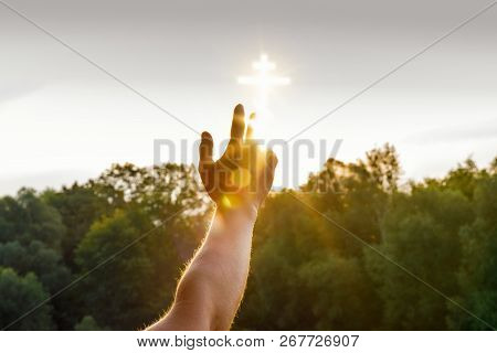 A Hand Is Reaching A Glowing Cross Image Which Is Situated In The Sky Above The Green Summer Trees. 