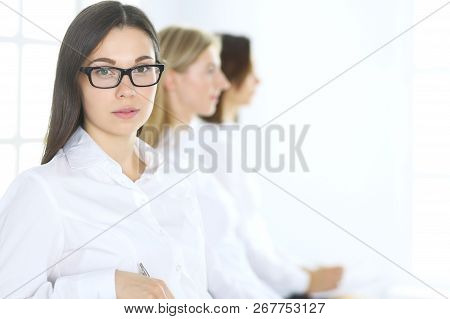 Attractive Businesswoman At Meeting Or Conference Against The Background Of Colleagues. Group Of Bus