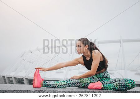 Picture Of Well-built Model Stratching Outside. She Sits And Leans Forward. Young Woman Is Serious A