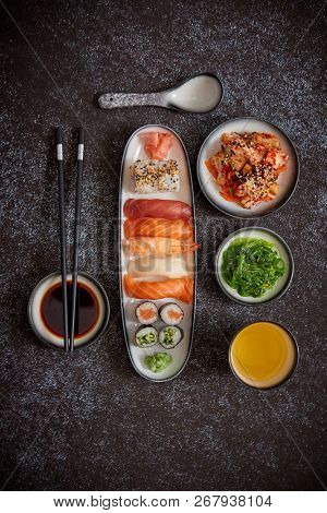Asian food assortment. Various sushi rolls placed on ceramic plates. Kimchi and goma wakame salads. Soy souce and chopsticks on sides. Grungy dark background with copy space.