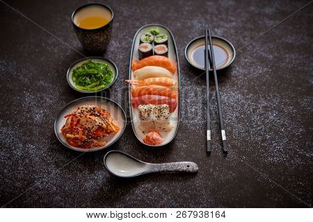 Asian food assortment. Various sushi rolls placed on ceramic plates. Kimchi and goma wakame salads. Soy souce and chopsticks on sides. Grungy dark background with copy space.