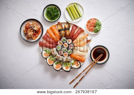 Asian food assortment. Various sushi rolls placed on round ceramic plate. Kimchi and goma wakame salads, fresh cucomber, ginger and wasabi. Soy souce and chopsticks on sides.