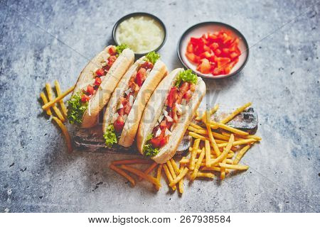Assortment of three tasety hot dogs, placed on wooden cutting board. Served with french fries, onion and tomato. View from above. Stone background.