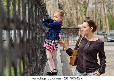 A Young Mother Plays With Her Daughter Blond Hair, Creeps On The Fence. The Concept Of The Family