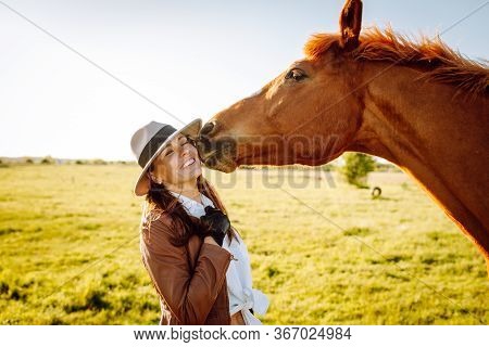 Beautiful Smiling Young Woman In A Hat And Gloves Playing With A Broun Horse In A Field On A Sunset.