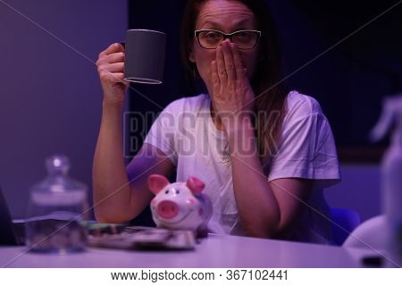Portrait Of Tired Young Woman Yawning Sitting In Room With Cup Of Coffee And Accounting Money From P