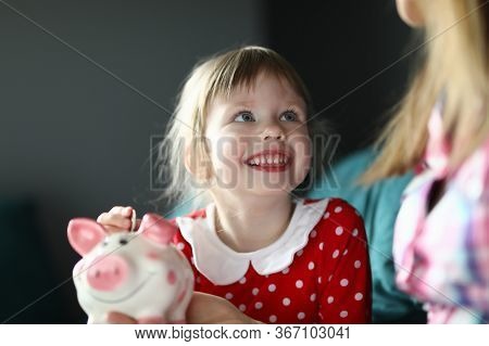 Portrait Of Smiling Cheerful Kid Putting Coin In Piggy Bank And Looking At Mother. Happy Joyful Chil