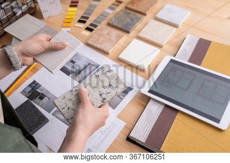 Hands of young female designer holding two samples of marble tile over wooden table with digital tablet, photos of home interior etc