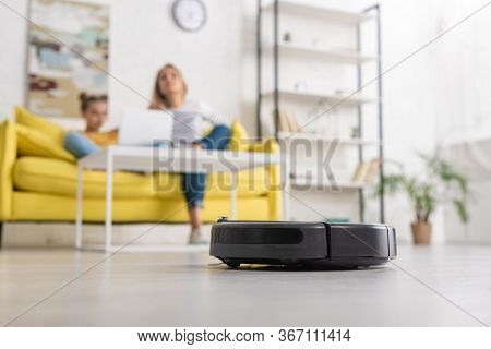 Selective Focus Of Robotic Vacuum Cleaner On Floor And Mother With Daughter On Sofa Near Coffee Tabl