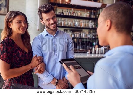 Couple Being Greeted By Maitre D Using Digital Tablet As They Arrive At Restaurant