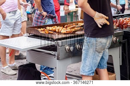 Young Man Holding Meat Shashlik In Tongs In Front Of A Huge Outdoor Grill, Hand Behind His Back, Pre