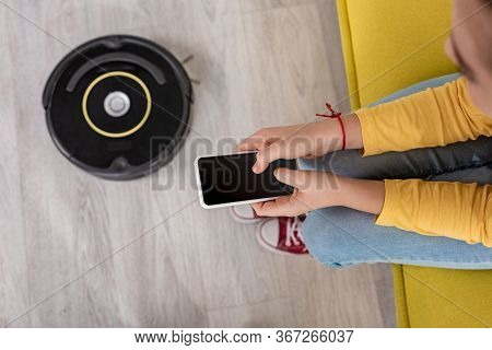 Cropped View Of Kid With Smartphone On Sofa Near Robotic Vacuum Cleaner On Floor In Living Room
