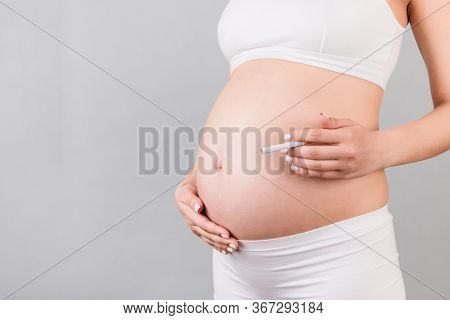 Close Up Of Pregnant Woman In White Underwear With A Cigarette Against Her Belly At Gray Background.