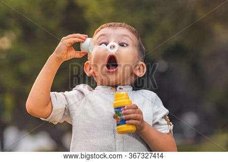 A Child With An Open Mouth And Big Eyes Blows Colorful Soap Bubbles In Nature.