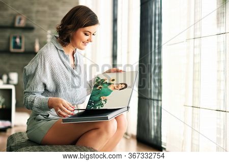 Young Happy Woamn Looking At Photo Album, Resting In Cozy Living Room