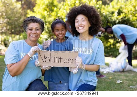 Mixed race girl spending time outside with her family, holding a volunteer sign with her mother and grandmother, looking at the camera and smiling, all wearing blue volunteer t shirts, on a sunny day