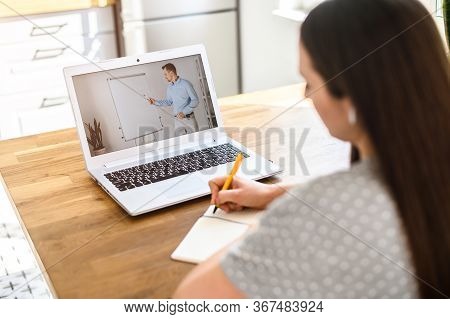 A Student Studies Online. A Student Girl Is Watching Online Classes And Writing A Syllabus In Notebo