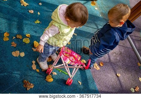 Little Cute Toddler Boy And A Beautiful Year-old Baby Girl In Blue Clothes Play In A Pink Toy Stroll