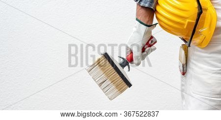 Caucasian House Painter Worker In White Work Overalls And With Protective Gloves, He Holds The Brush