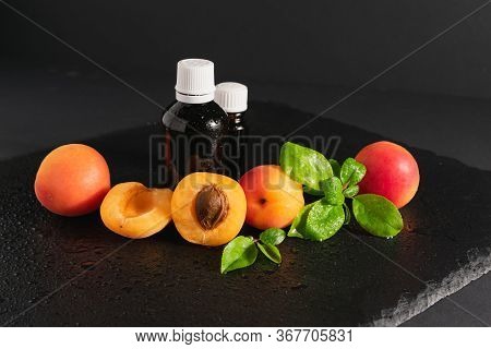 Dark Glass Bottles With Apricot Kernel Oil And Ripe Fresh Apricots On A Black Stone Background.