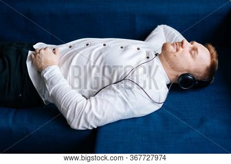 Young Photogenic Man Is Resting On Blue Soft Sofa With Headphones At Home Listening To Relaxing Musi