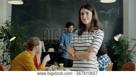 Portrait Of Pretty Young Cheerful Girl Turning Face To Camera And Smiling. Team Of Startuppers On Ba