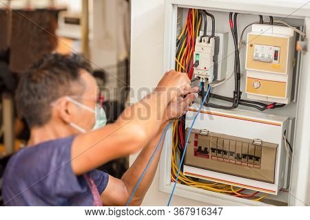 Electrician With Medical Mask Engineer Works With Electric Cable Wires Of Fuse Switch Box.