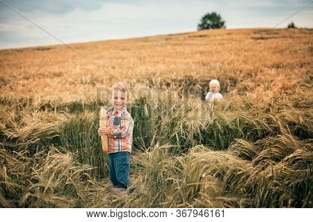 A Blond Boy In An Orange Plaid Shirt And Green Trousers Stands In A Rye Field, Smiles And Holds A Wh