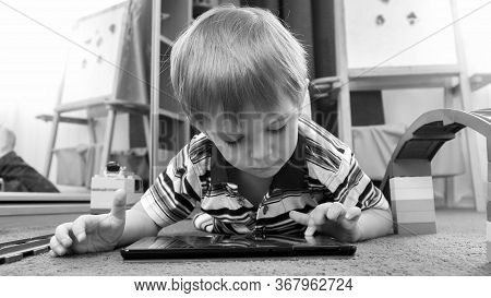 Portrait Of Cute Little Boy Playing Games On Tablet Lying On Carpet At Living Room