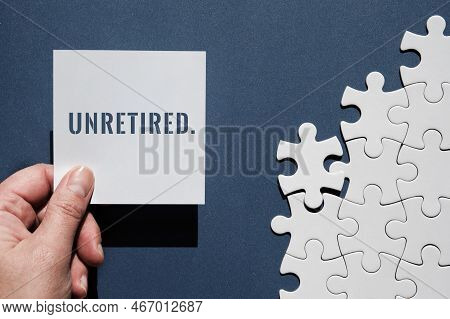 Unretired Word Written On Square Paper Note In Hand. Top View, Flat Lay On Dark Blue Monochromatic P