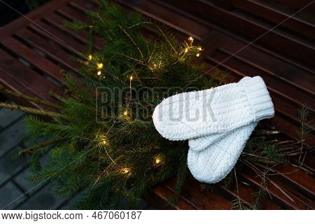 Branches Of A Coniferous Tree Decorated With A Garland Lie On A Bench Along With Knitted White Mitte