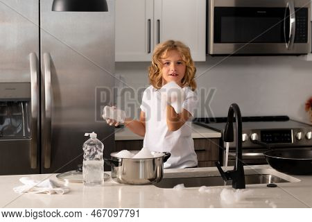 Kid Boy Washing Dishes In The Kitchen Interior. Child With Sponge With Dish Washing Liquid Is Doing 