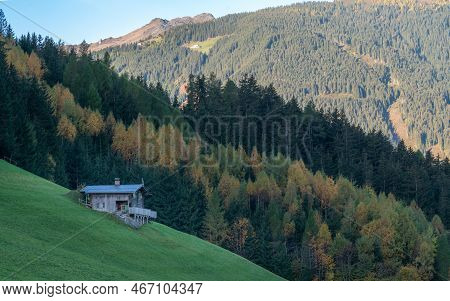Rustic Wooden Cabin On A Steep Slope Of Mountain Meadow In Austrial Alps With Sun Lit Alpine Peak In
