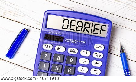Flat Lay Or Top View Of Blue Pen And Calculator With Debrief Text On Light Wooden Table.