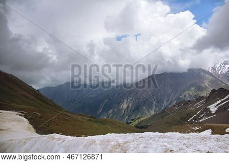 Mountain Range With Peaks, Thick Clouds On The Peaks, Sunlight On The Slopes, Snow In The Foreground