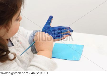 Child Paints Her Own Hand With Blue Paint And Paintbrush Getting Messy And Having Fun.